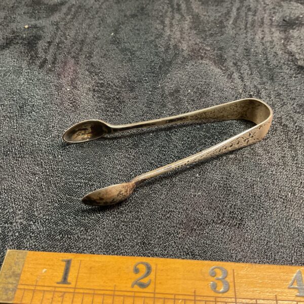 Antique silver sugar tongs on ruler for scale.