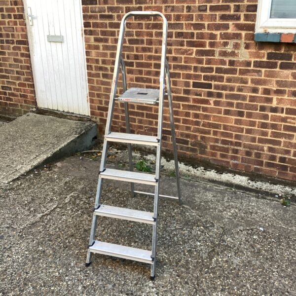 Front view of a three-step aluminium ladder against a brick wall.