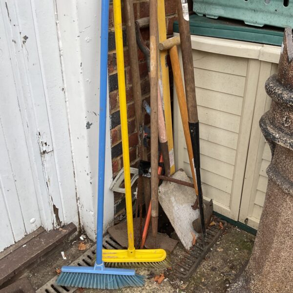 Bundle of garden tools including brooms and shovels leaning against a wall.