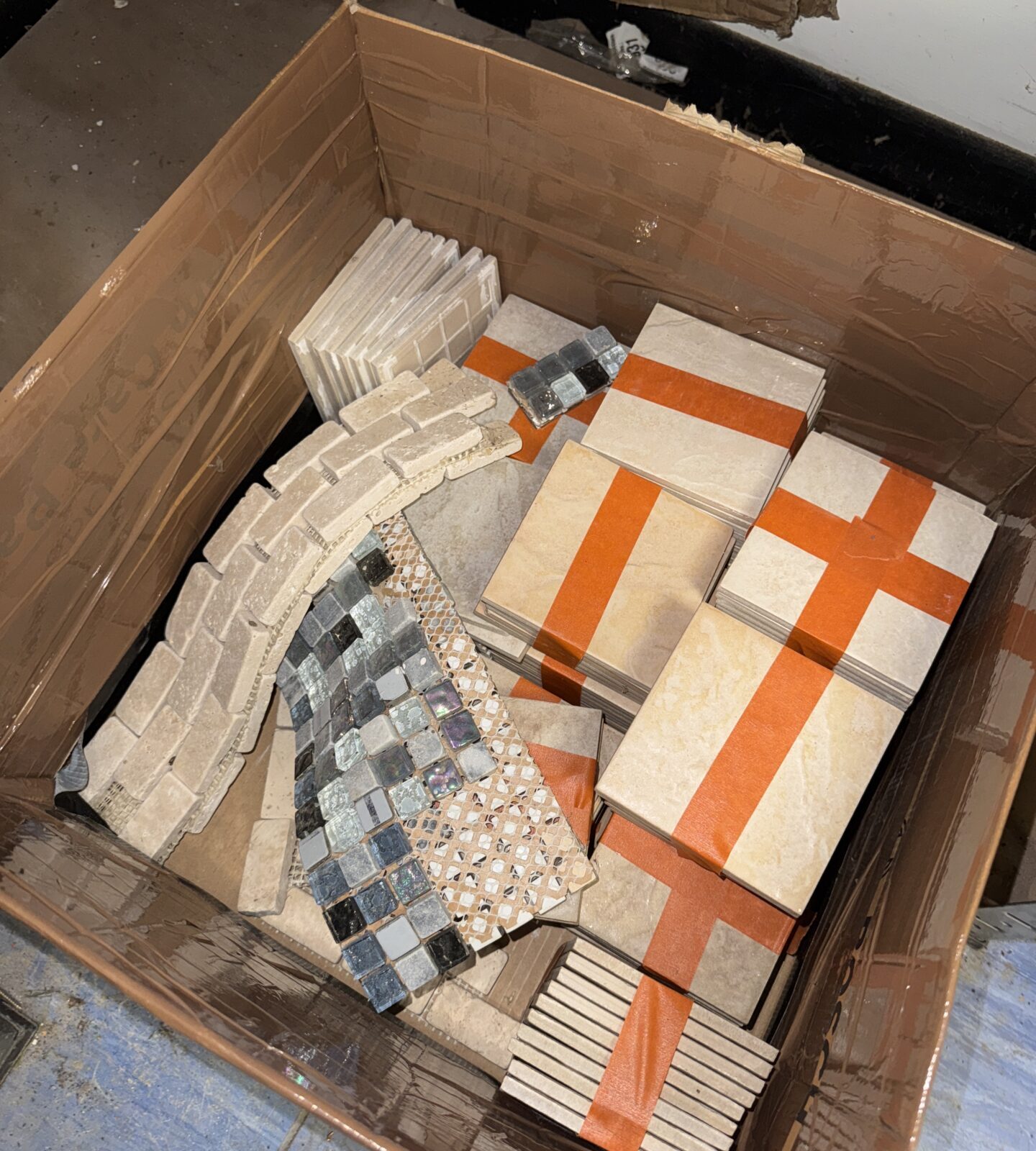Mixed mosaic and square tile samples in a cardboard box.