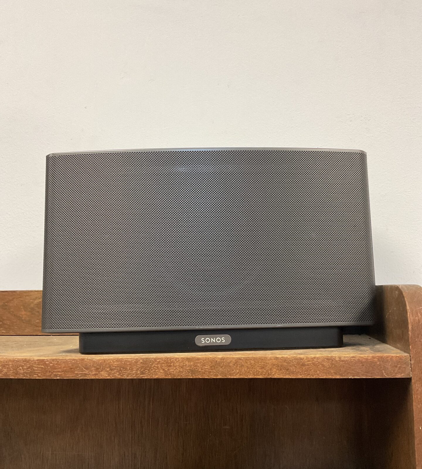 Black Sonos Play 5 wireless speaker on a wooden shelf.
