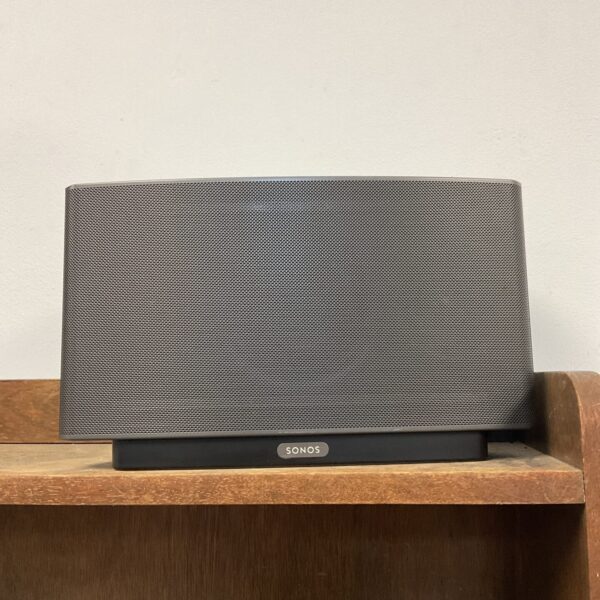Black Sonos Play 5 wireless speaker on a wooden shelf.