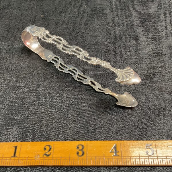 Ornate antique silver sugar tongs on dark fabric.