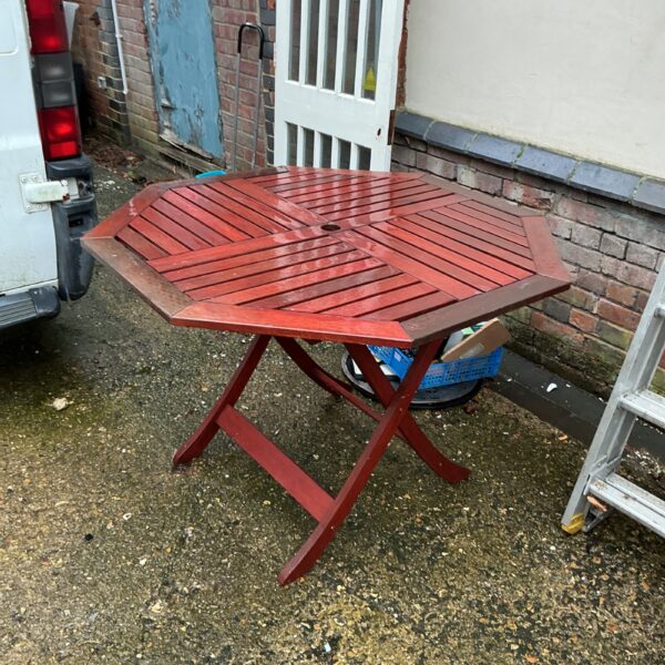 Reddish-brown teak garden table with folding legs in a garden setting.