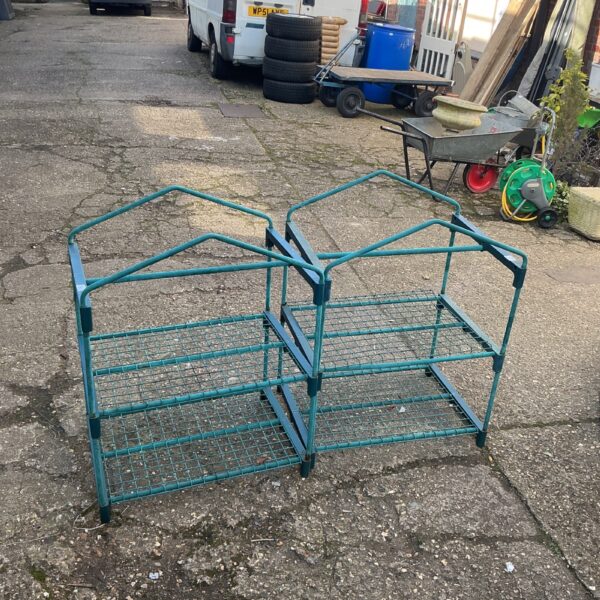 Two green metal tiered garden plant stands in a garden setting.