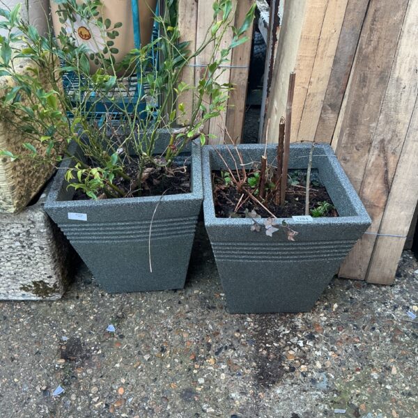 Two grey garden pots with green plants in a garden setting.