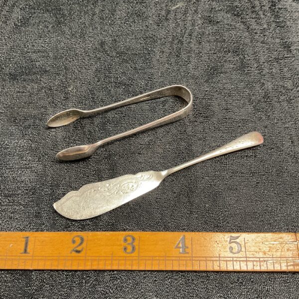 Vintage silver sugar tongs and fish knife set on grey fabric.