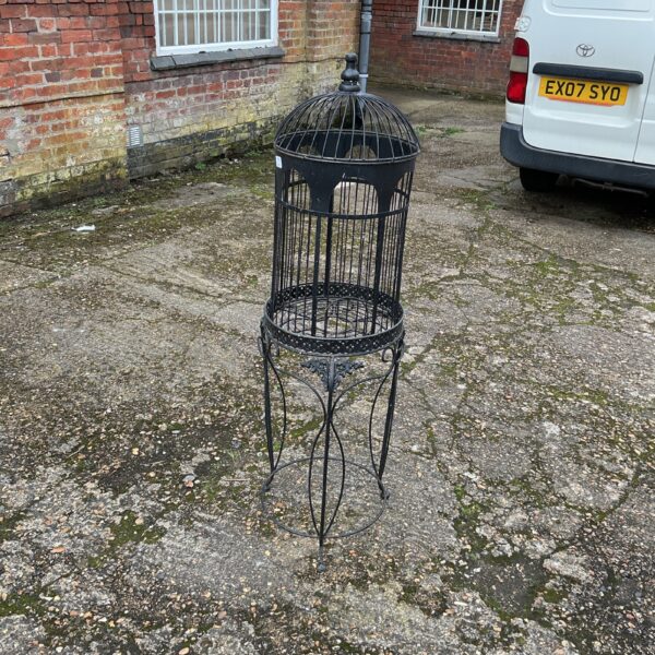 Ornate vintage metal bird cage on stand in garden setting.
