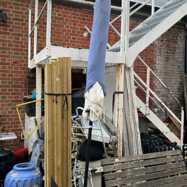 Large garden parasol with weighted base in a cluttered garden setting.