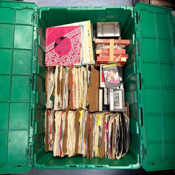 Large lot of mixed 7 inch vinyl records in a green crate.