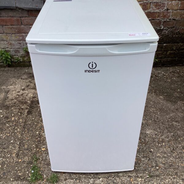 White Indesit fridge standing against a brick wall.