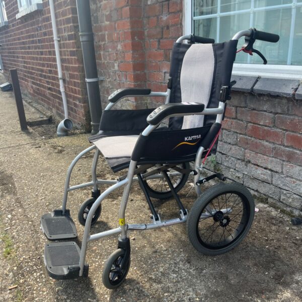 Silver Karma Ergo Lite transit wheelchair against brick wall.
