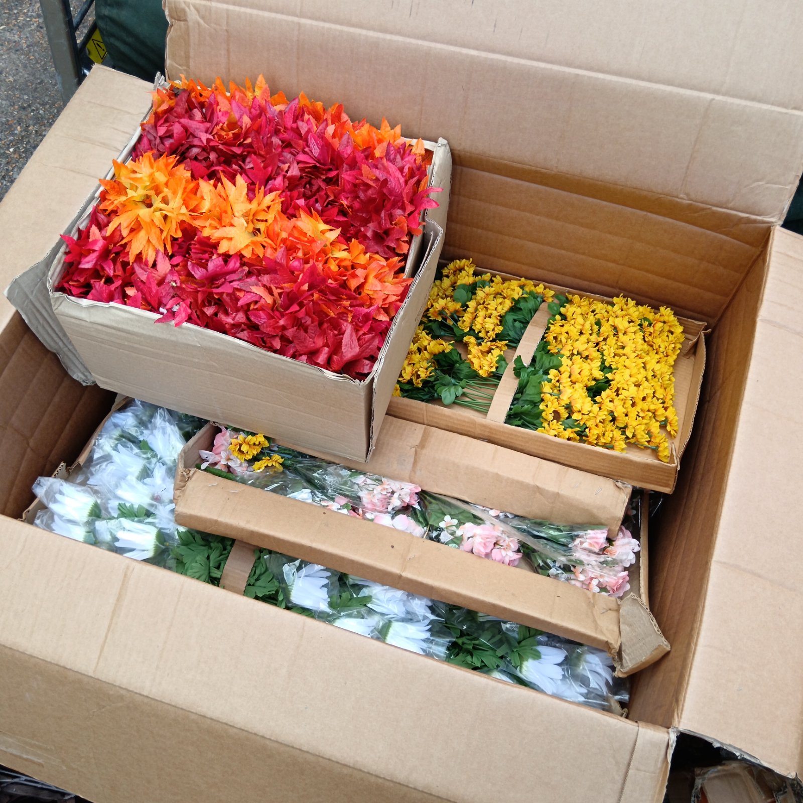 Bulk lot of colourful faux flowers in a cardboard box.