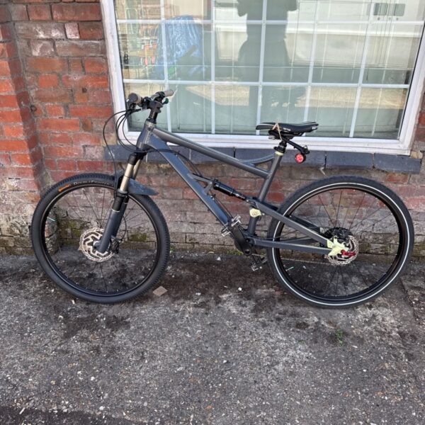 Grey full suspension mountain bike leaning against brick wall.