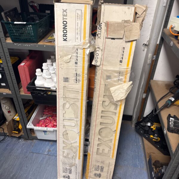 Stack of Kronotex Exquisit laminate flooring boxes in a workshop.