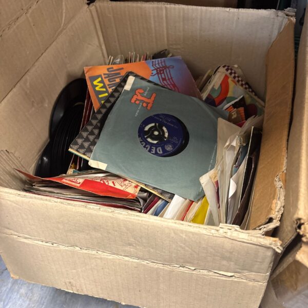 Large lot of mixed 7 inch vinyl records in a cardboard box.
