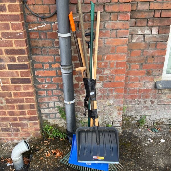 Bundle of garden tools including rakes and pitchforks leaning against a wall.