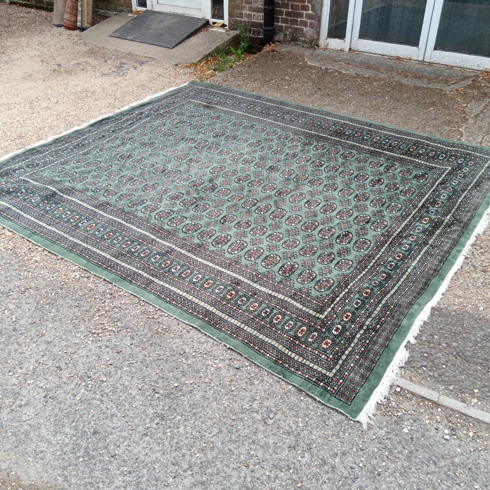 Green Pakistan Bokhara rug with geometric patterns displayed on a driveway.