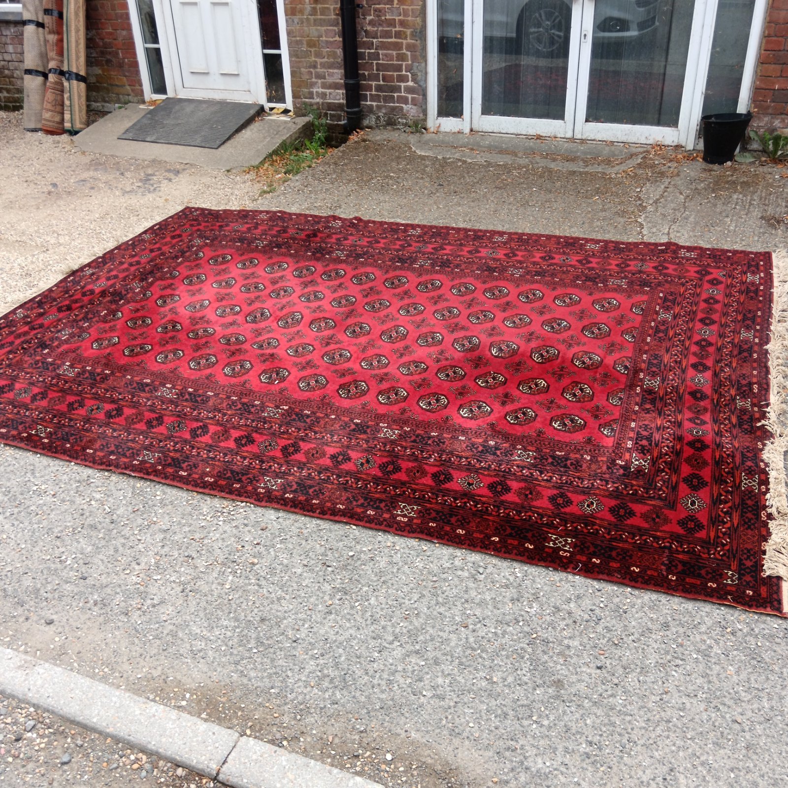 Red Louis de Poortere Mossoul rug displayed on a gravel driveway.