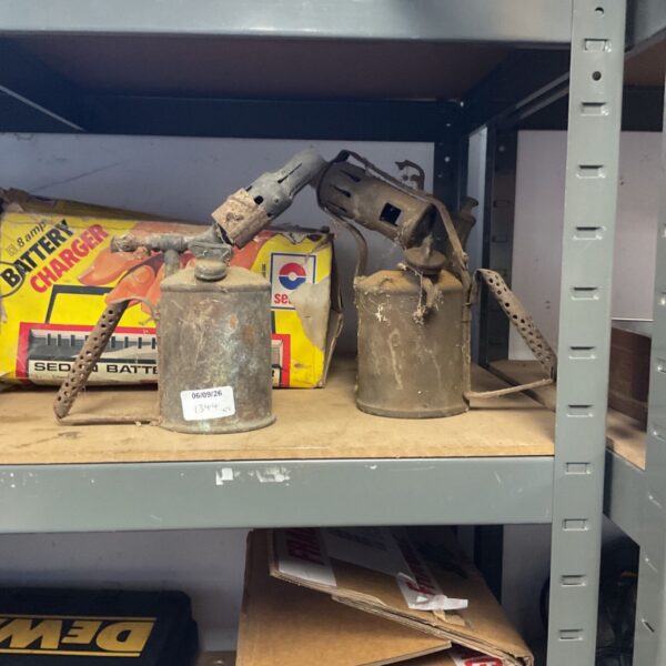 Two vintage brass paraffin blowtorches displayed against a backdrop.