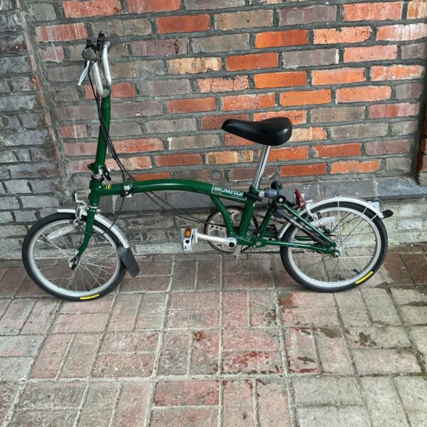Green Brompton folding bicycle standing against a brick wall.