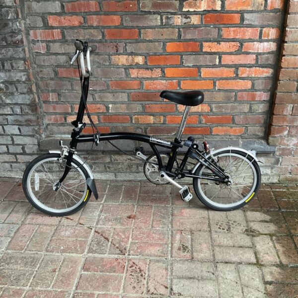 Black Brompton folding bicycle leaning against brick wall.