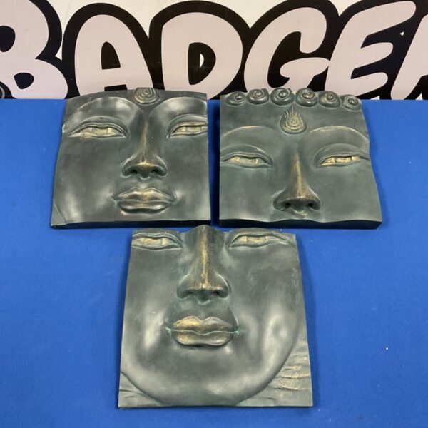 Set of three green bronze-effect Buddha face wall panels on blue surface
