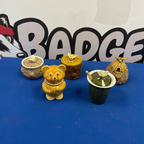Five glazed ceramic honey pots including bear and beehive designs on blue surface