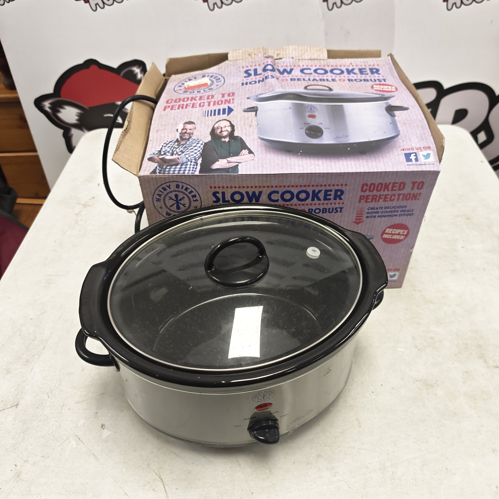 Hairy Bikers stainless steel slow cooker with glass lid beside its printed box