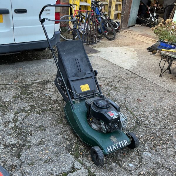 Green Hayter petrol lawn mower with black grass box on a paved yard