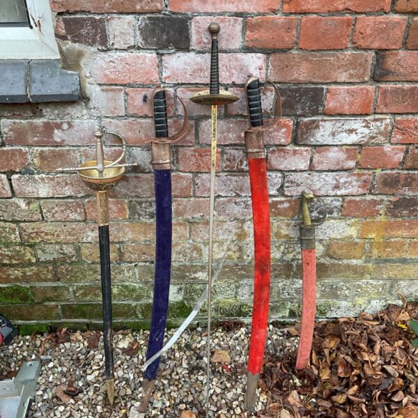 Five ceremonial swords with brass hilts and coloured scabbards leaning against a brick wall