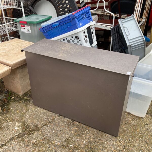 Brown metal garden storage box with overhanging lid on paved ground