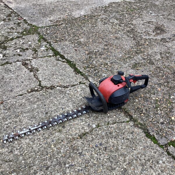 Red and black petrol hedge cutter with long double-edged blade resting on paving