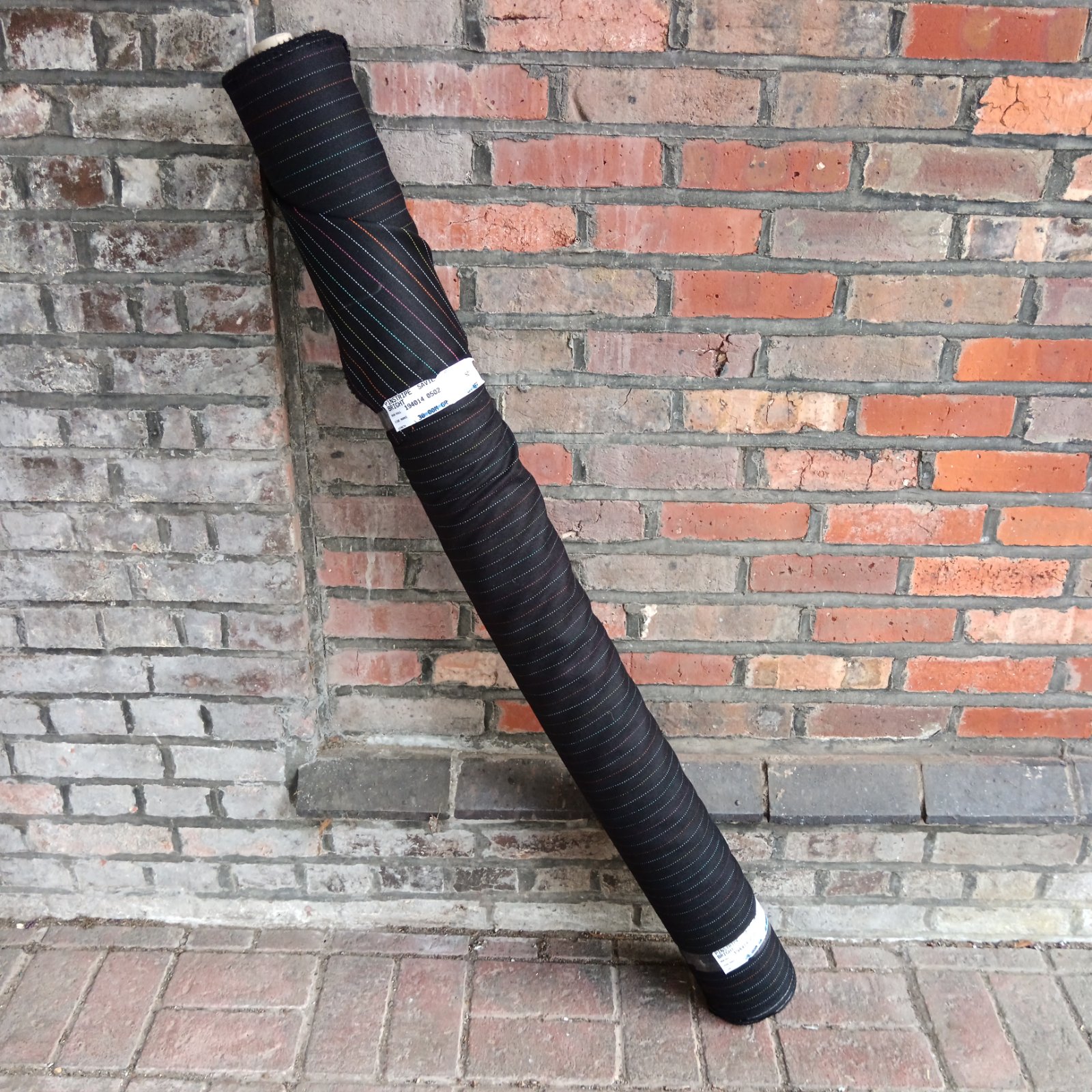 Large roll of dark pinstripe Savile Row suit fabric against brick wall.