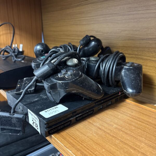 Black Sony PlayStation 2 Slim console with several controllers and cables on shelf