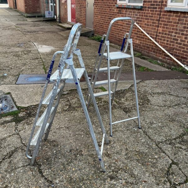 Two aluminium step ladders with blue grip handles outdoors on paved ground