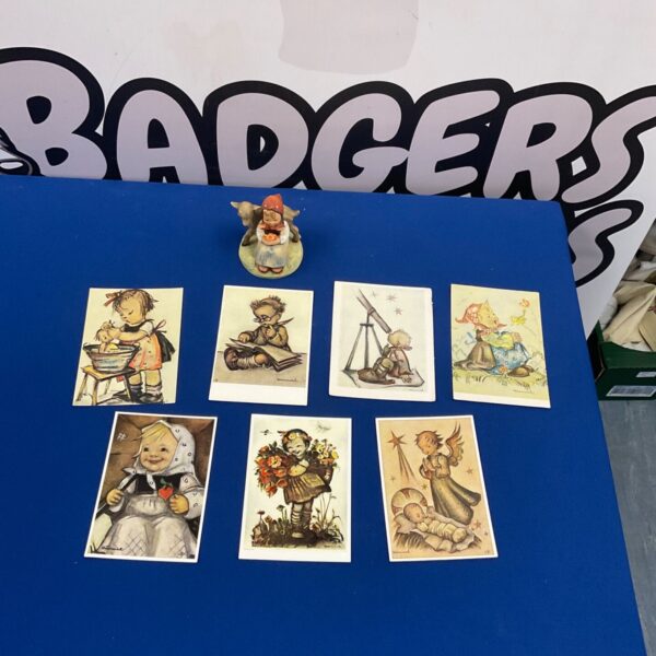 Vintage Hummel Goebel figurine beside seven German Hummel postcards on blue surface