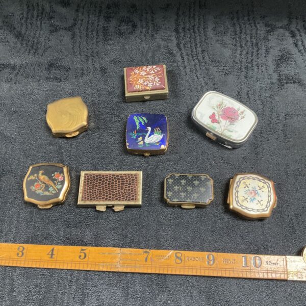 Eight assorted vintage pill boxes with enamel, gilt and printed designs on a dark background