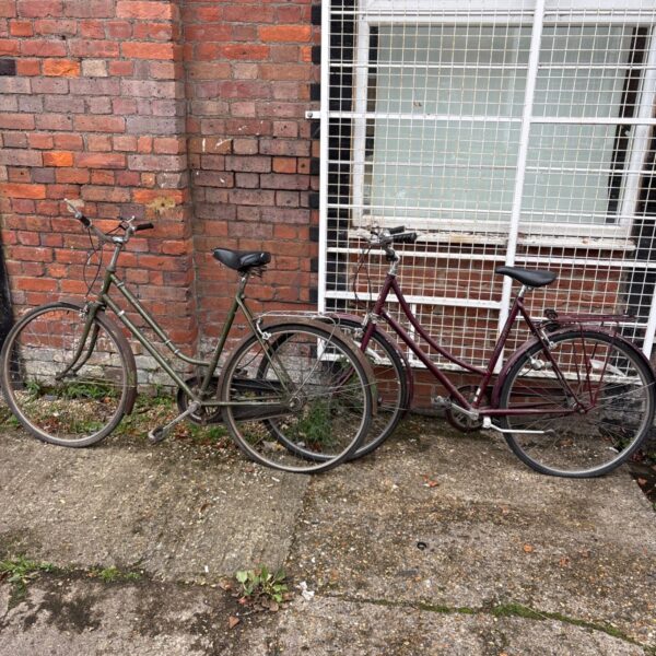 Two vintage Raleigh bicycles, olive green and maroon, parked against a brick wall